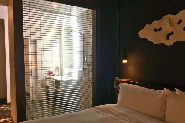 Chambre d'hôtel avec lit blanc, lumière douce filtrant par la fenêtre et par la porte entrouverte de la salle de bain