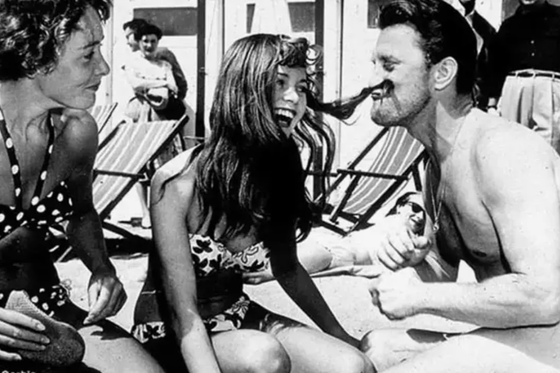Brigitte Bardot et Kirk Douglas discutant sur le sable, souriants.